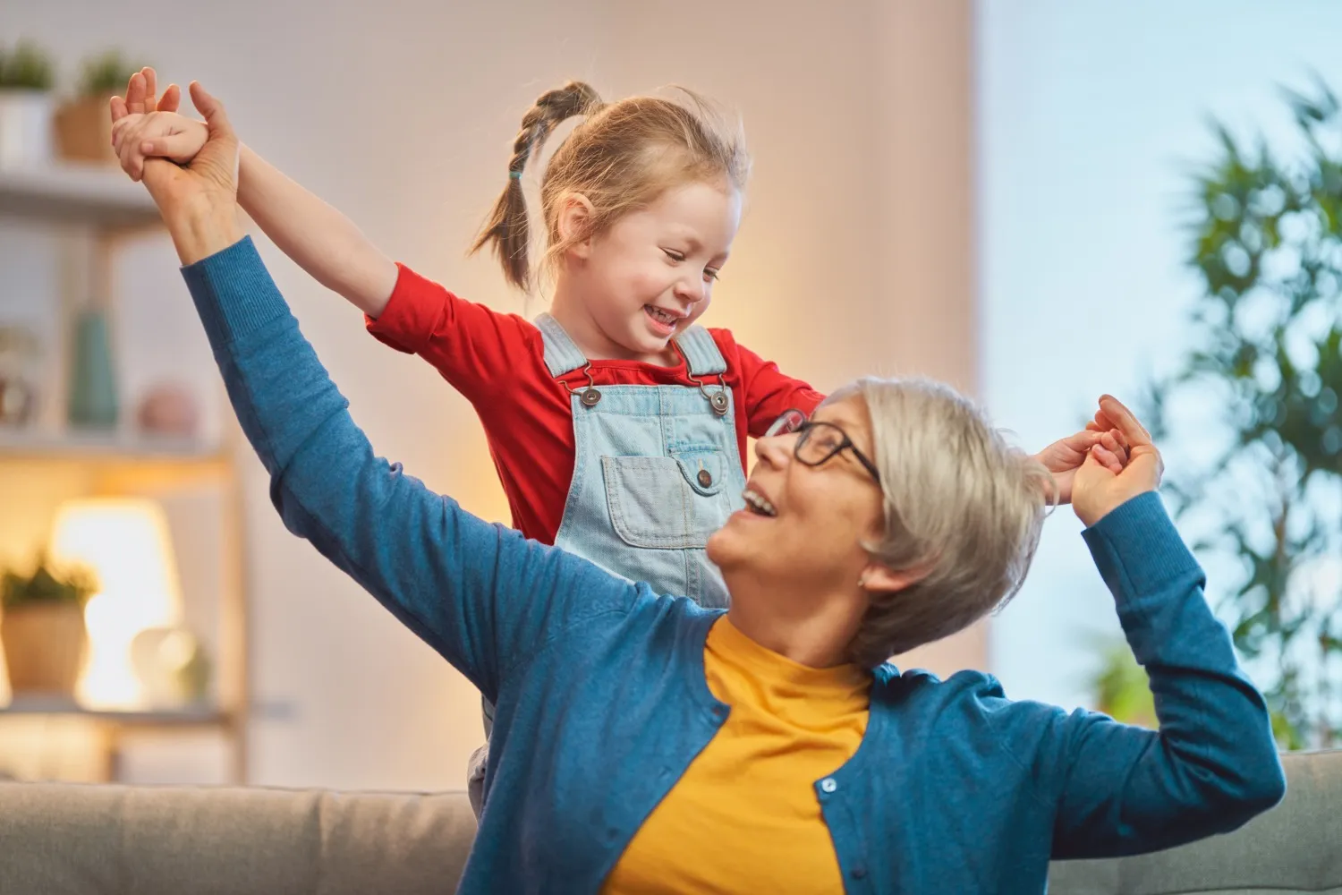 Smiling elderly woman with glasses playing and holding hands with a joyful young girl in a red shirt and denim overalls indoors.