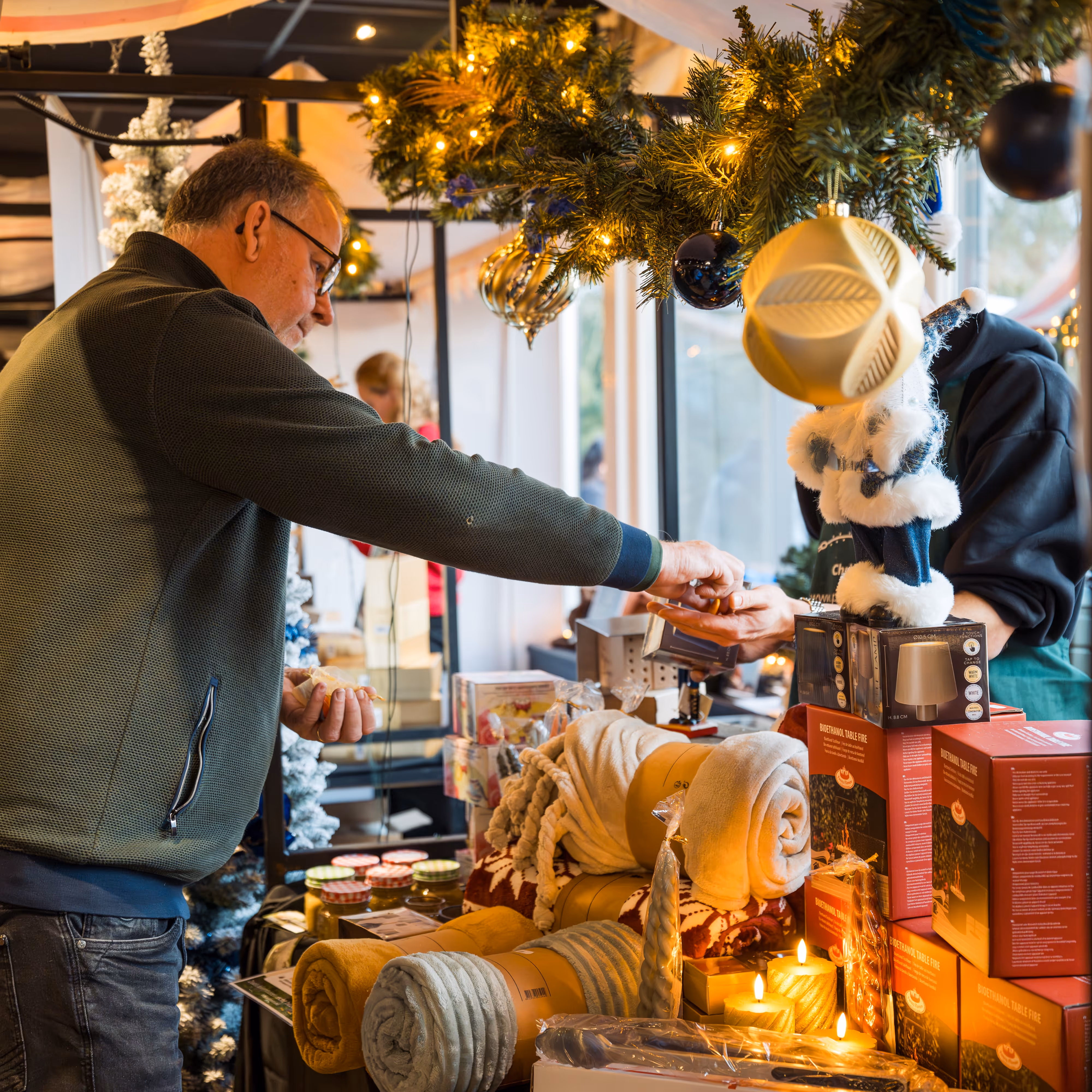Man die met muntjes een betaling doet bij een kerstmarkt kraam met kerstdecoraties en geschenken zoals dekens en kaarsen.