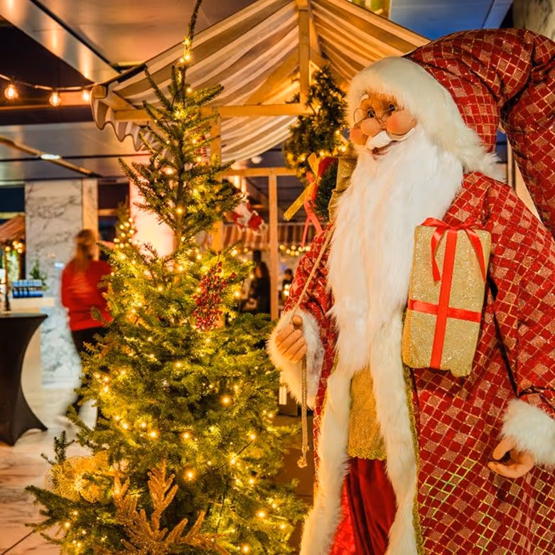 Grote kerstmanpop met rood gewaad en geschenk naast verlichte kerstboom op sfeervolle kerstmarkt.