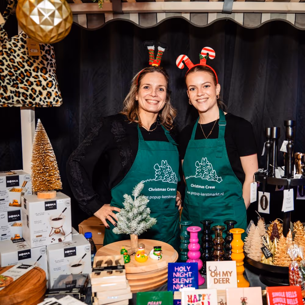 Twee lachende vrouwen met groene kerstschorten van Pop-up Kerstmarkt staan achter een kerstkraam met kerstdecoraties en cadeaus voor het kerstpakket