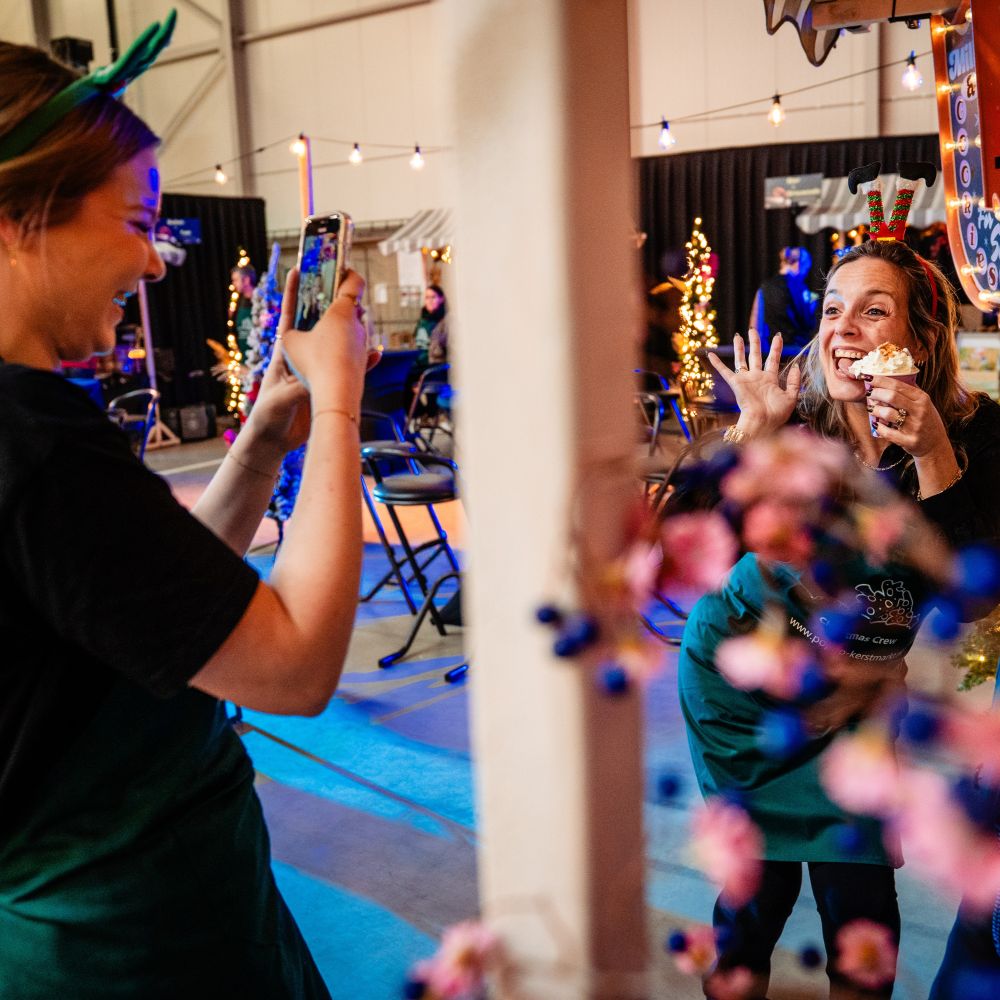 Vrouw neemt tijdens opbouw kerstmarkt foto van lachende medewerkster die een dessert vasthoudt en naar de camera zwaait op een feestelijke kerstmarkt.