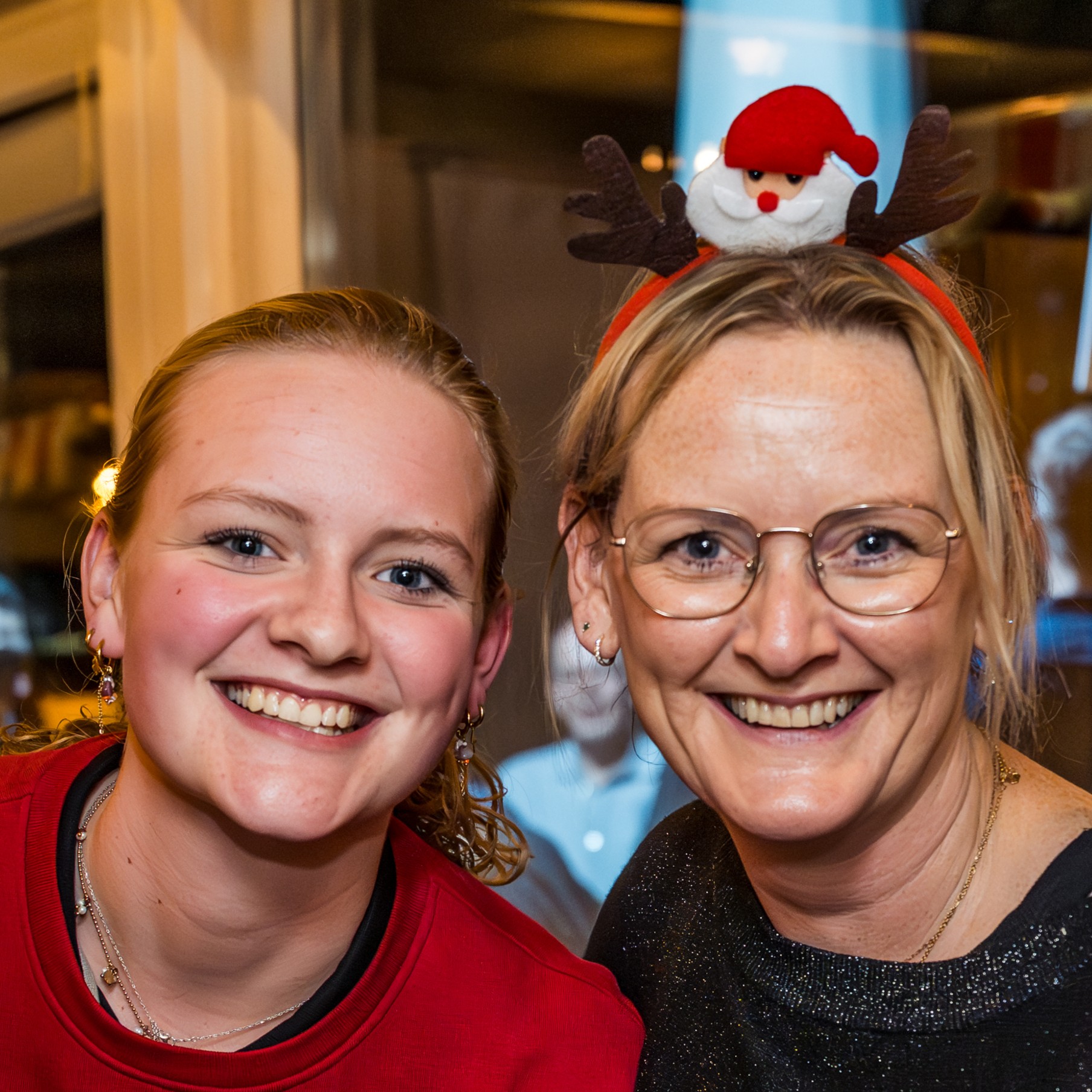 Twee lachende medewerkers dicht bij elkaar, de een draagt een rood shirt, de ander draagt een haarband met een kerstman en rendiergewei op een kerstmarkt op locatie.