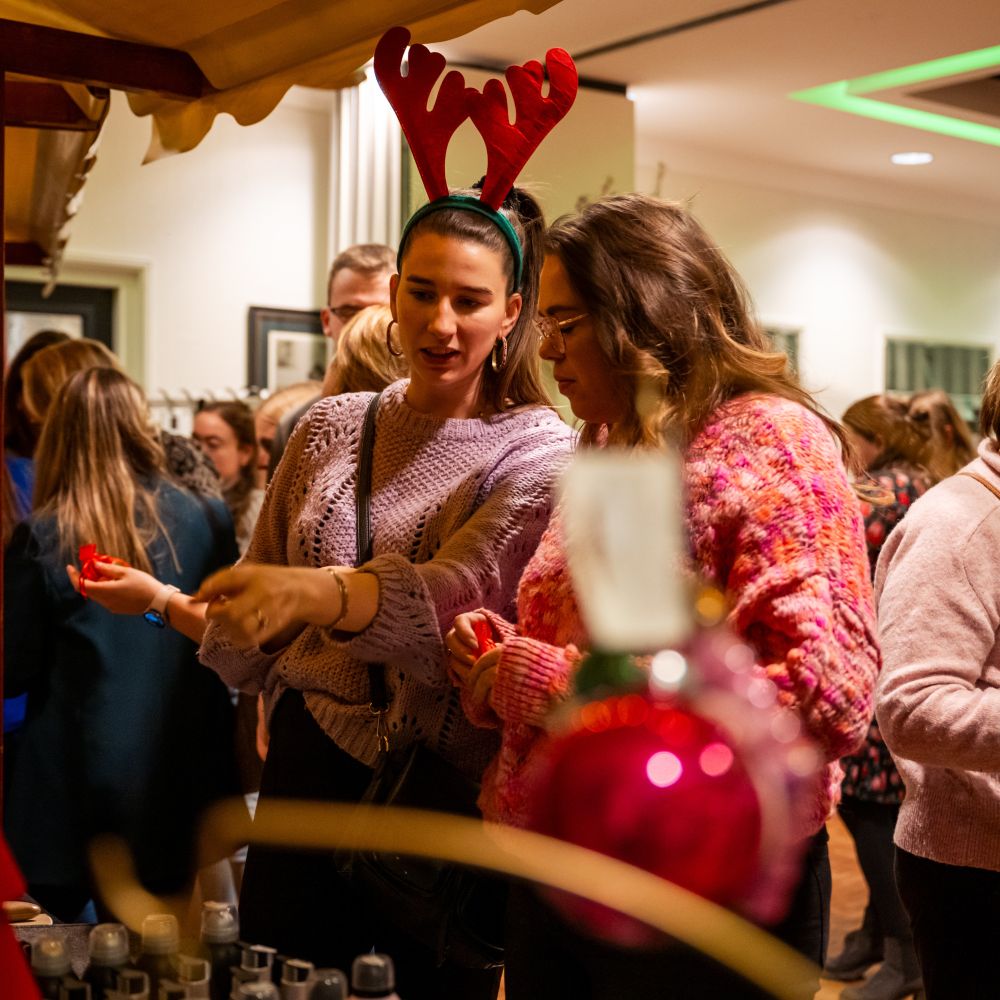 Twee medewerkers van de Pop-up kerstmarkt, een man met een kerstmuts en groene schort en de eventmanager met een kerstaccessoire en bril, poseren samen bij een versierde kerstboom en een tafel met spellen in een gezellige bar.