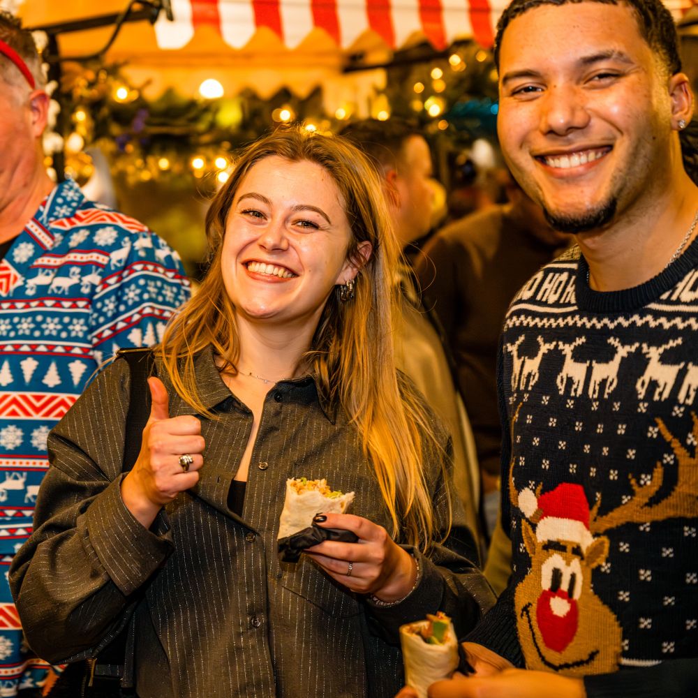 Mensen ruiken aan verpakte geschenken op een kerstevenement met sfeerverlichting op de achtergrond.