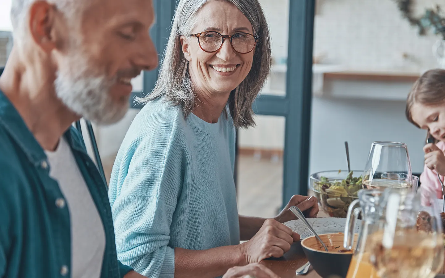 Smiling older woman wearing glasses enjoying a family meal, representing improved quality of life with hearing