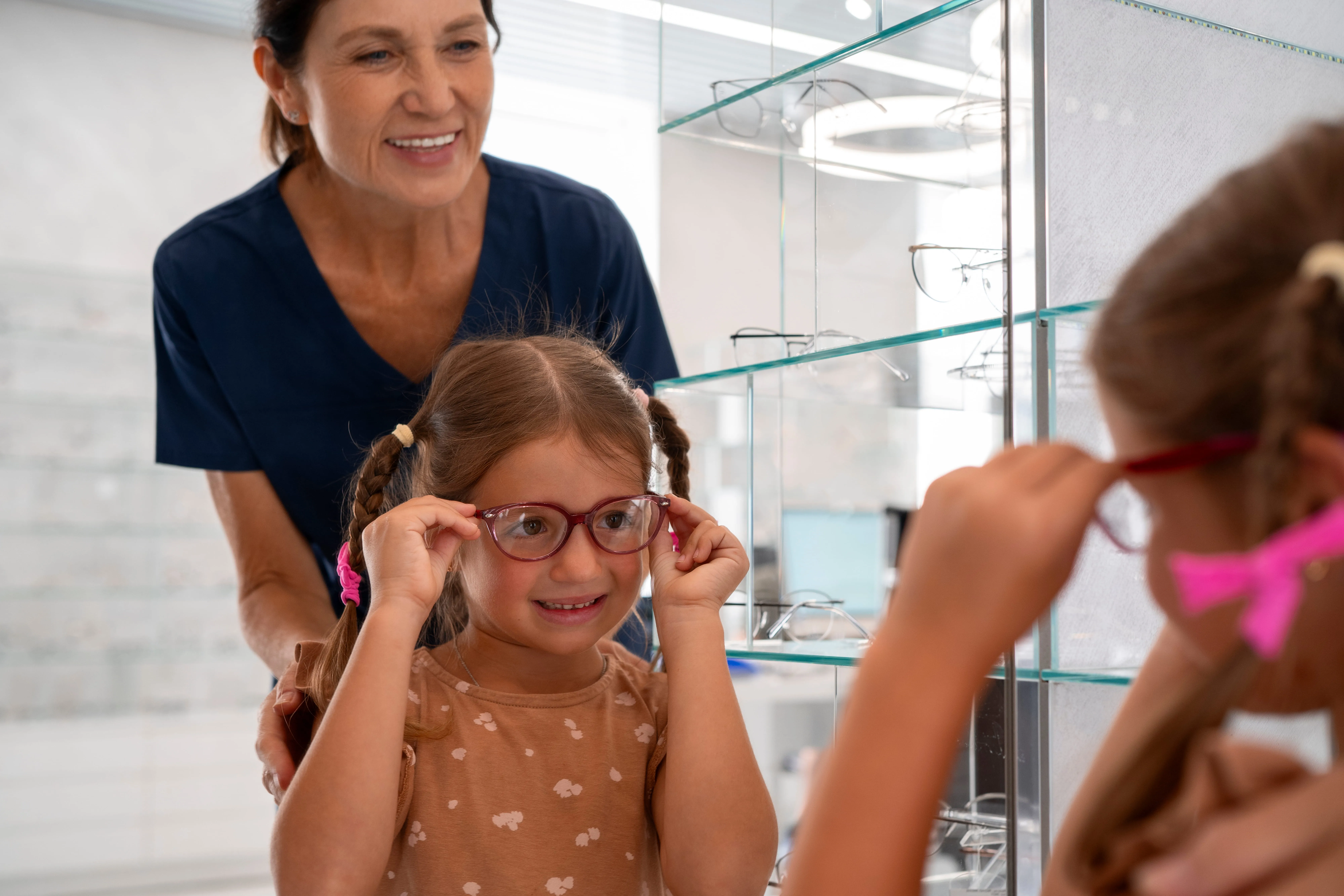 Young girl tries on glasses at optometrist with smiling adult supervisor nearby