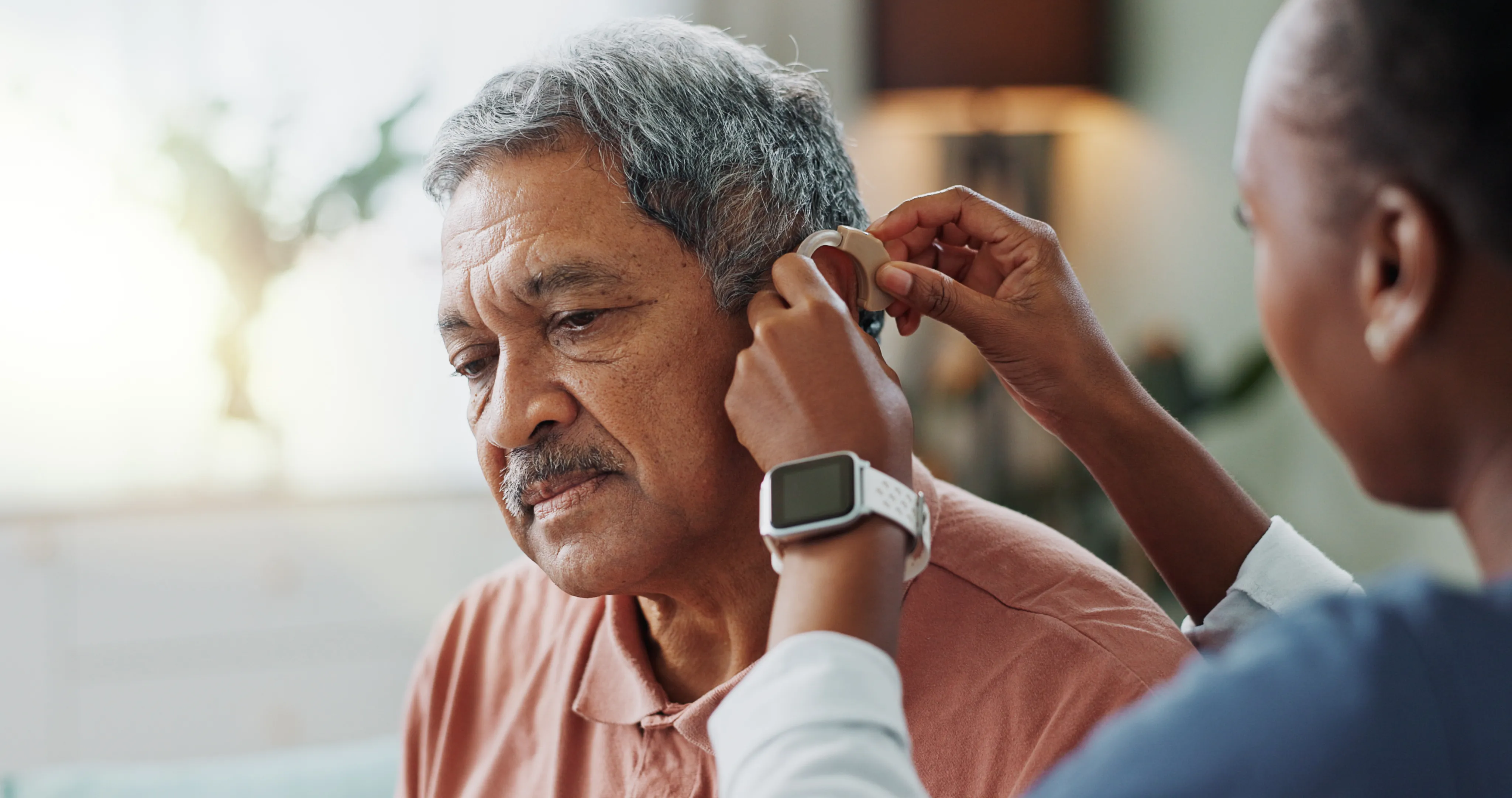 Senior man adjusting hearing aid in ear, assisted by another person indoors