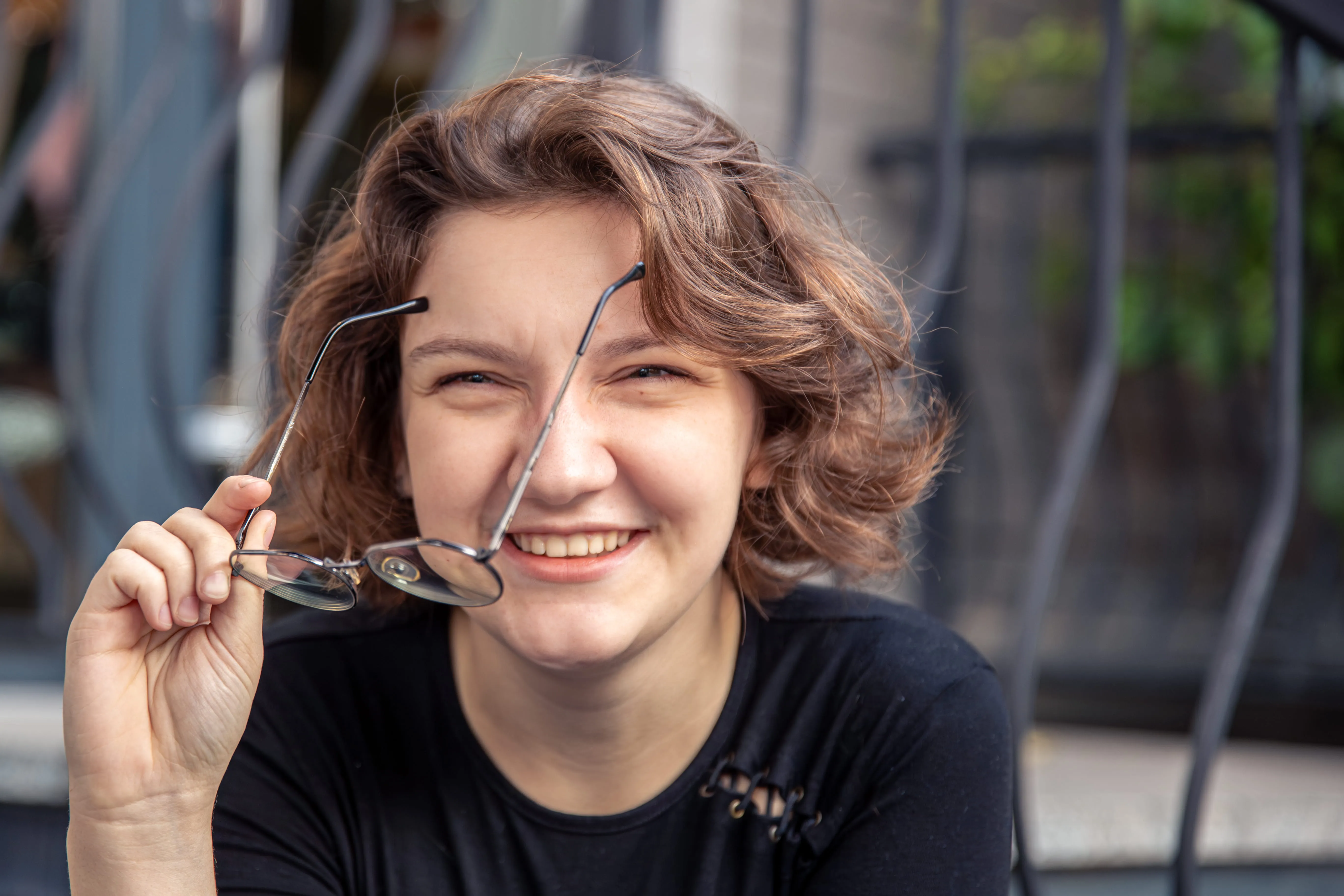 Smiling woman with wavy brown hair holding glasses in hand outdoors