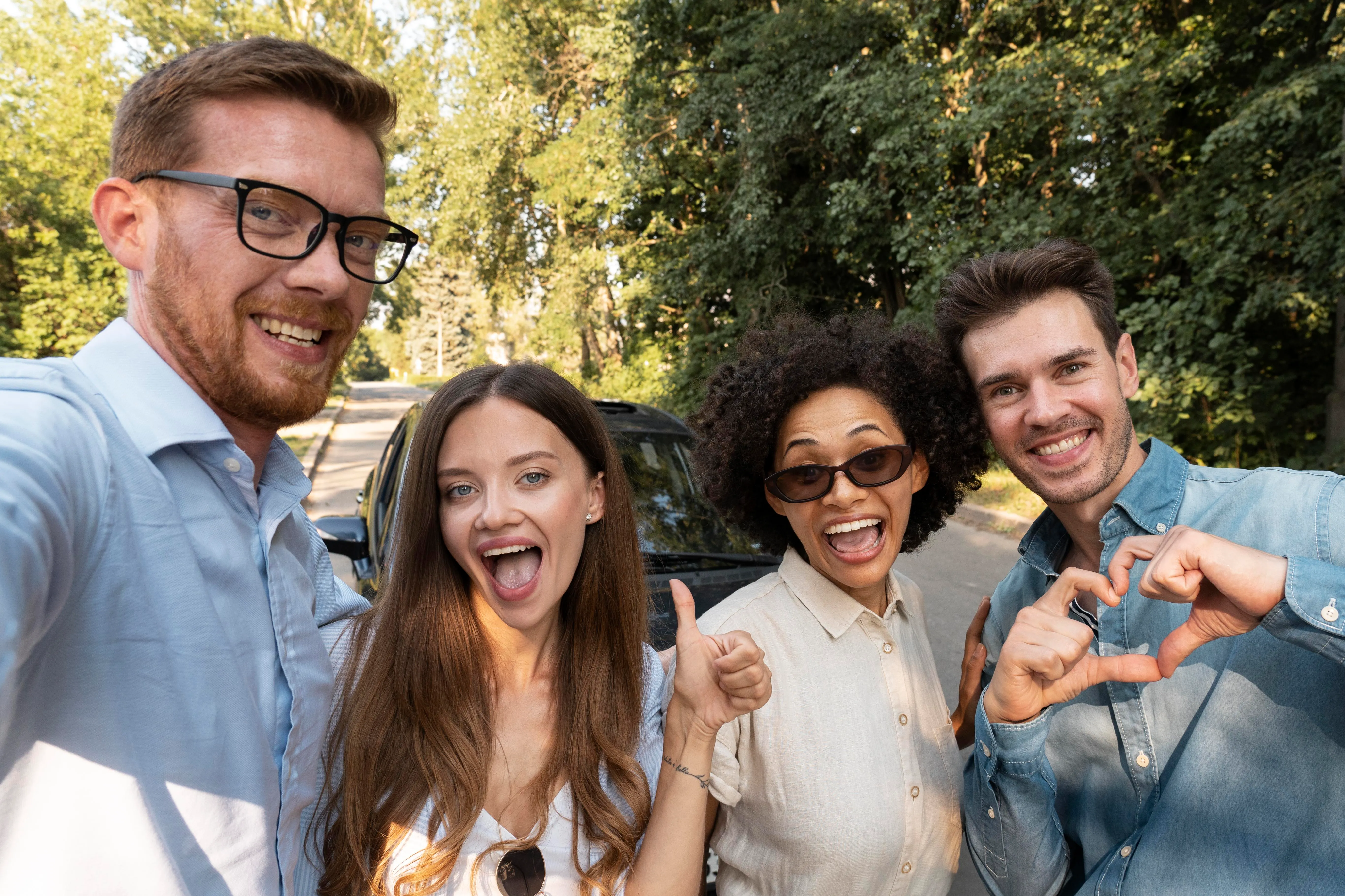 Four happy friends making hand gestures outdoors near a car and trees.