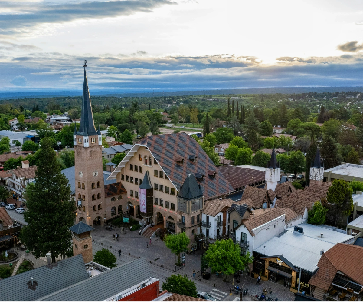Aerial view of a town square with a tall clock tower, European-style buildings, and surrounding greenery under a cloudy sky.