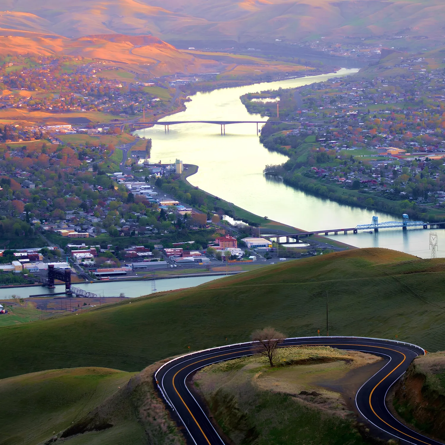 Curved road winding through green hills leading to a river with multiple bridges over a town during sunset.