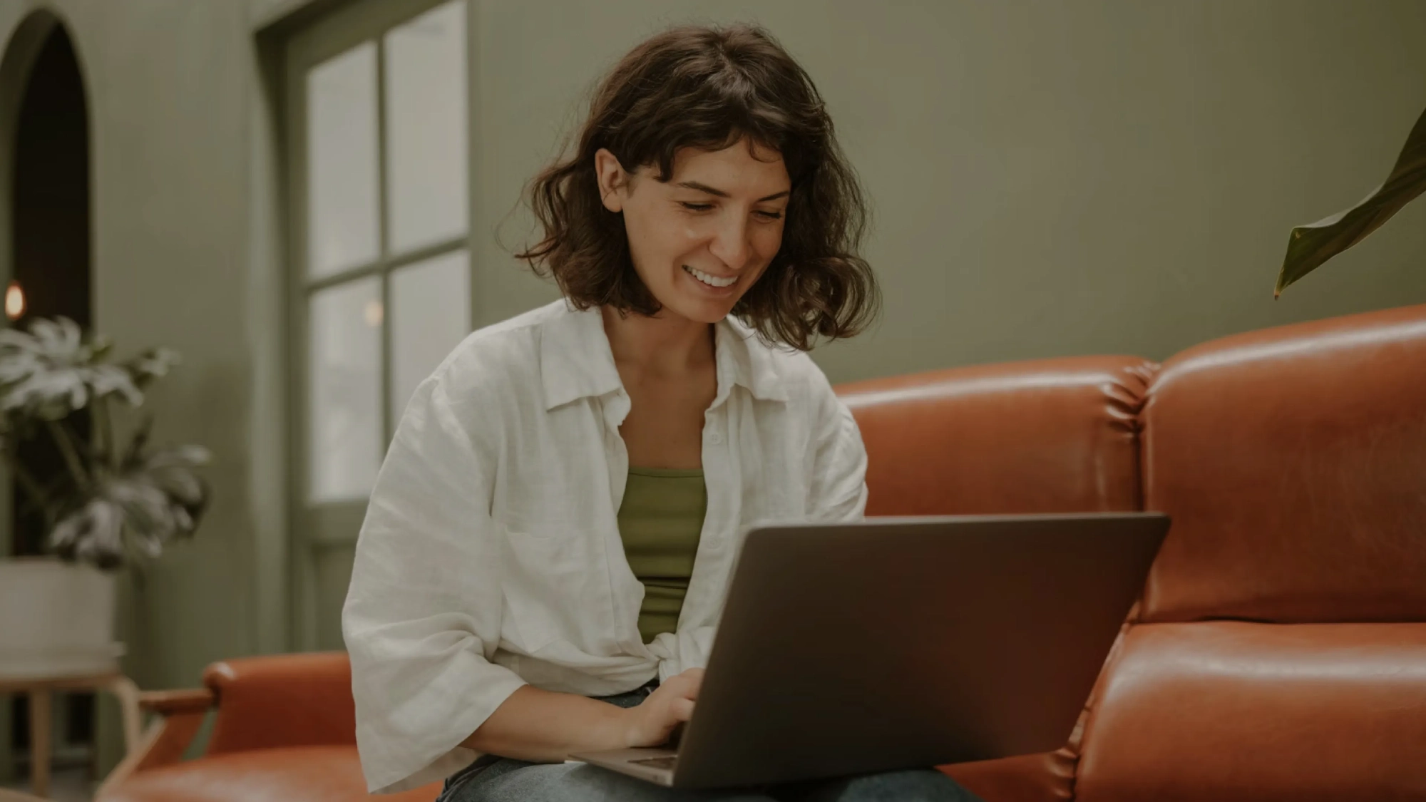 Smiling woman with curly brown hair sitting on a brown leather couch working on a laptop.