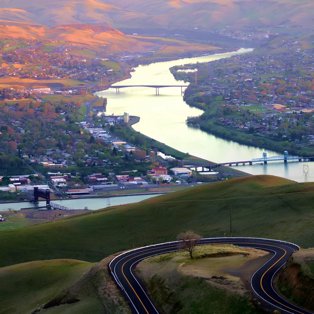 Curved road winding through green hills leading to a river with multiple bridges over a town during sunset.