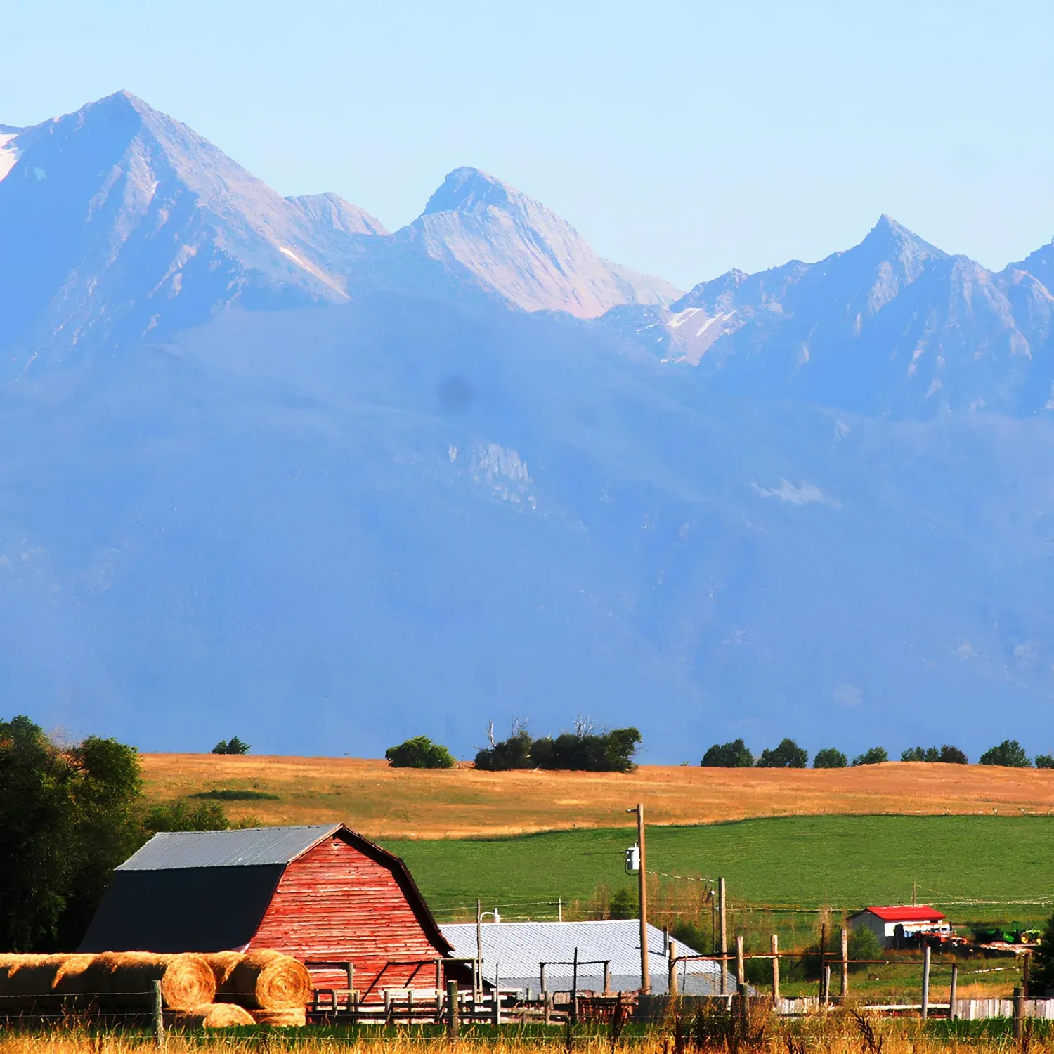 Red barn with hay bales in front of green and golden fields, set against a backdrop of tall blue mountains under clear sky.