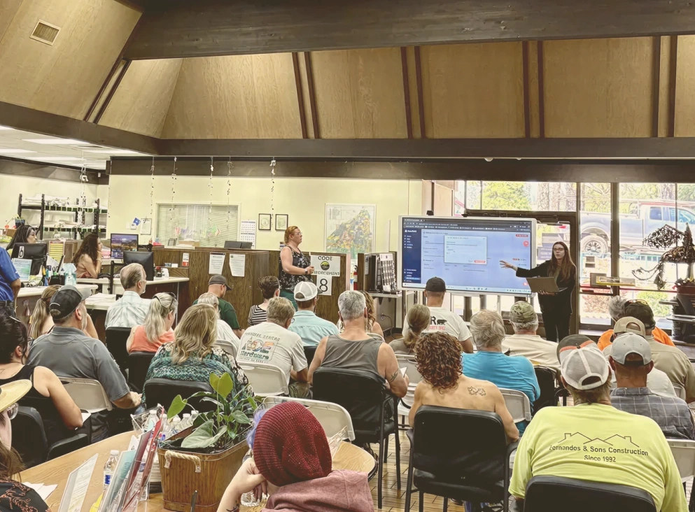 Group of adults seated and listening to a woman presenting information on a large monitor in an office setting.