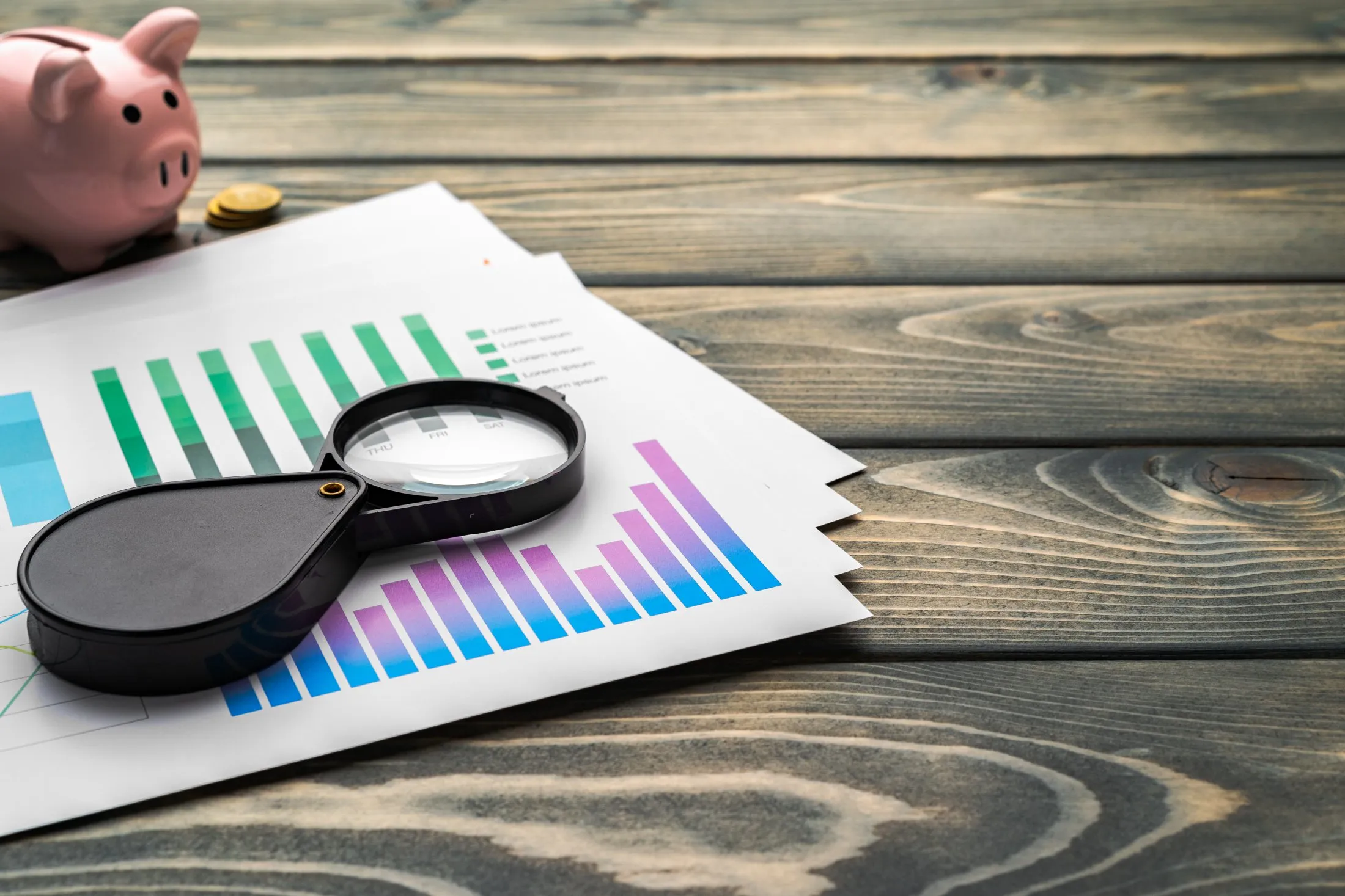 Magnifying glass on colorful bar graph papers with a pink piggy bank and coins on wood table.
