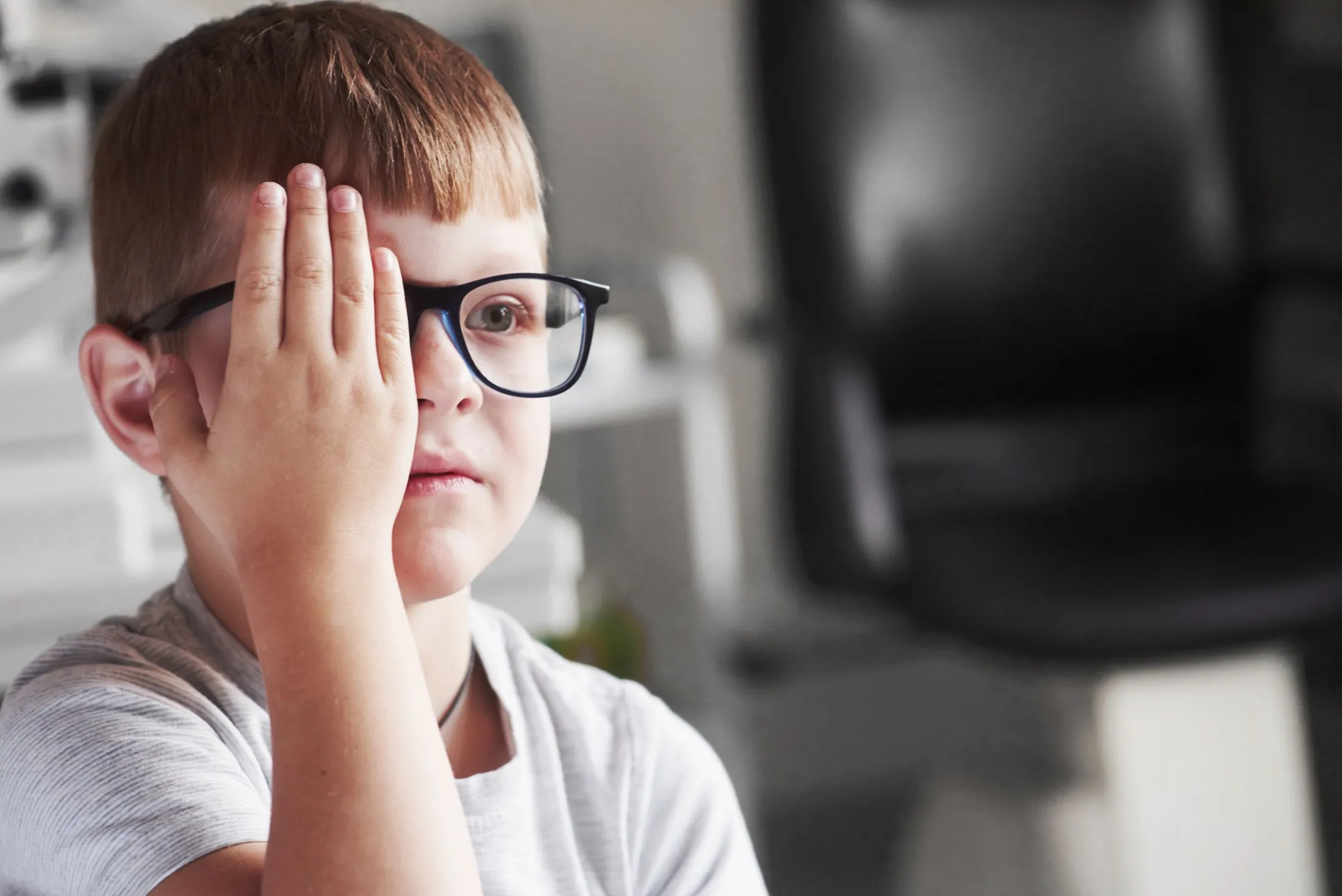 Young boy wearing glasses covering his right eye with his hand, focused on something ahead.