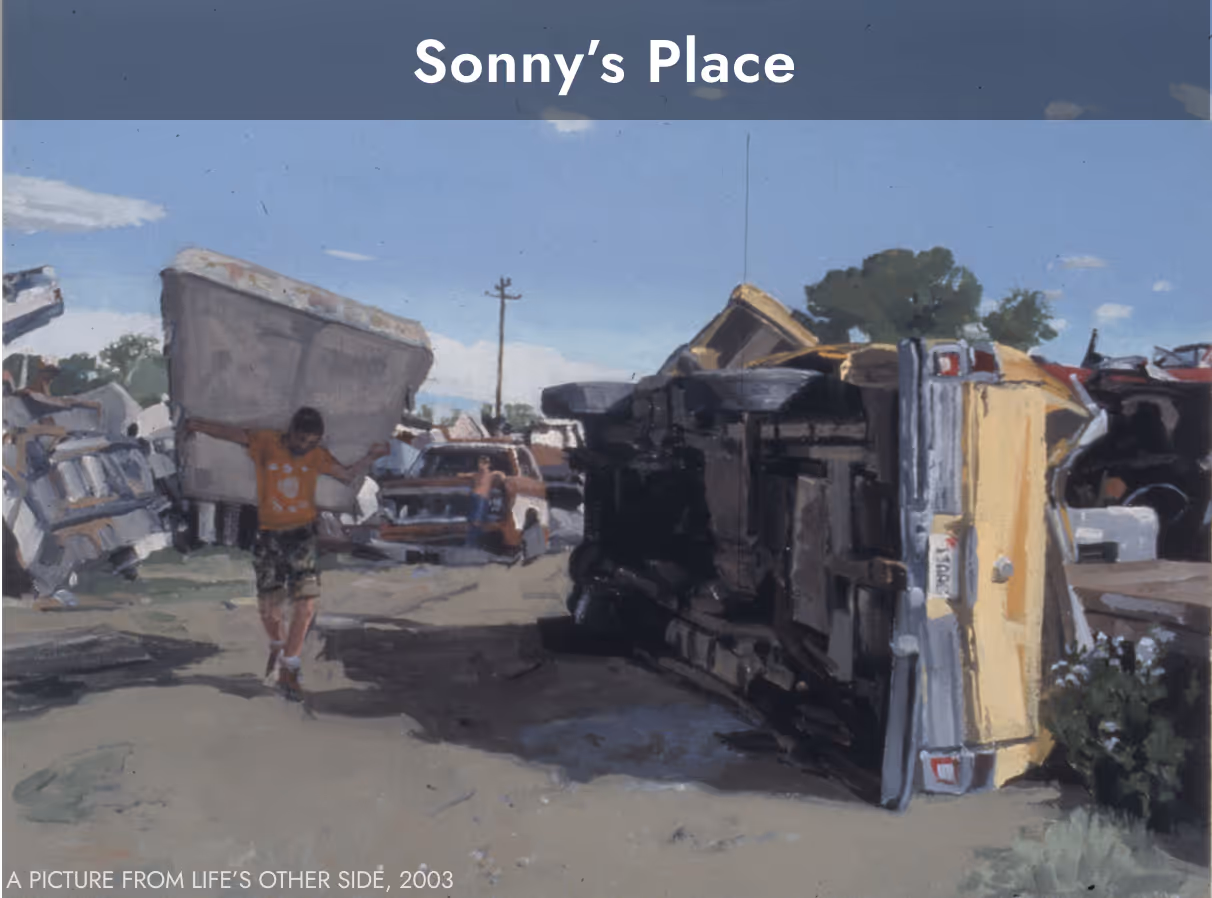 A man in a junkyard carrying a mattress on his back next to a vehicle on its side.