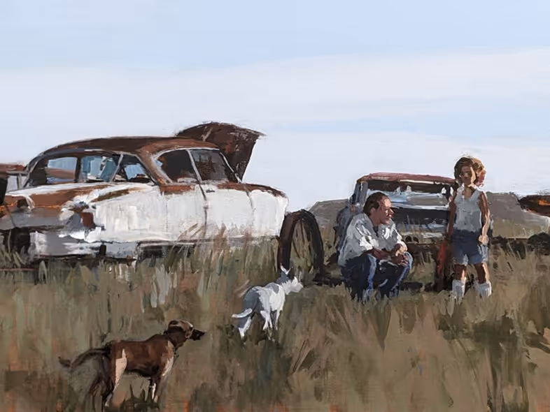 A man crouching and a girl standing near two dogs and old cars in a grassy field.