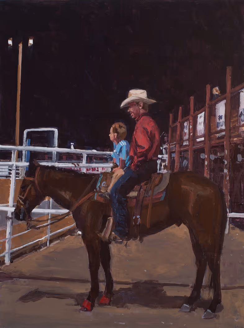 Man in cowboy hat and child sitting on horse in fenced arena at night.