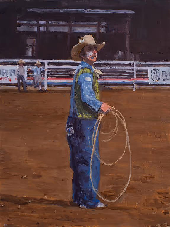 Rodeo clown in cowboy hat holding a lasso on dirt arena with fence and spectators in background.