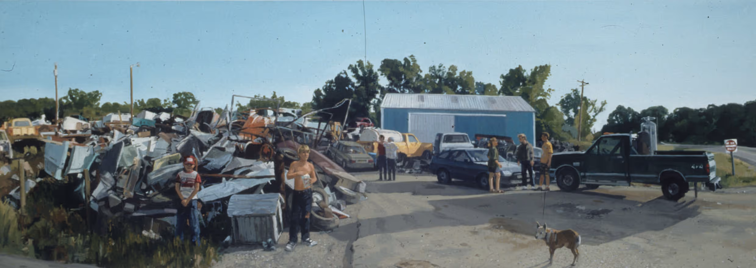 Group of children and adults near junkyard piles, cars, a blue shed, truck on a sunny day.