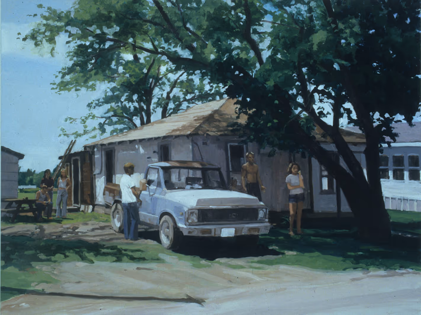 People gathered outside a weathered house with a blue pickup truck under tall trees on a sunny day.