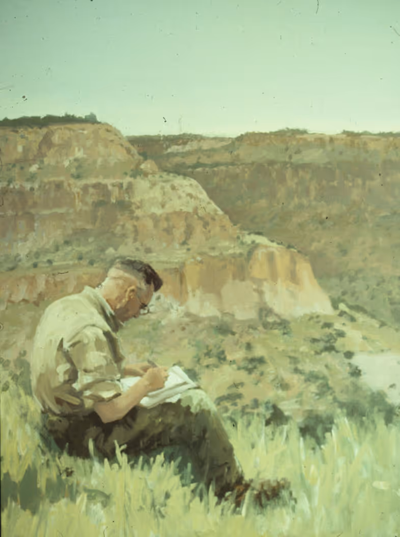 Man sitting in grass sketching or writing with canyon cliffs in the background.