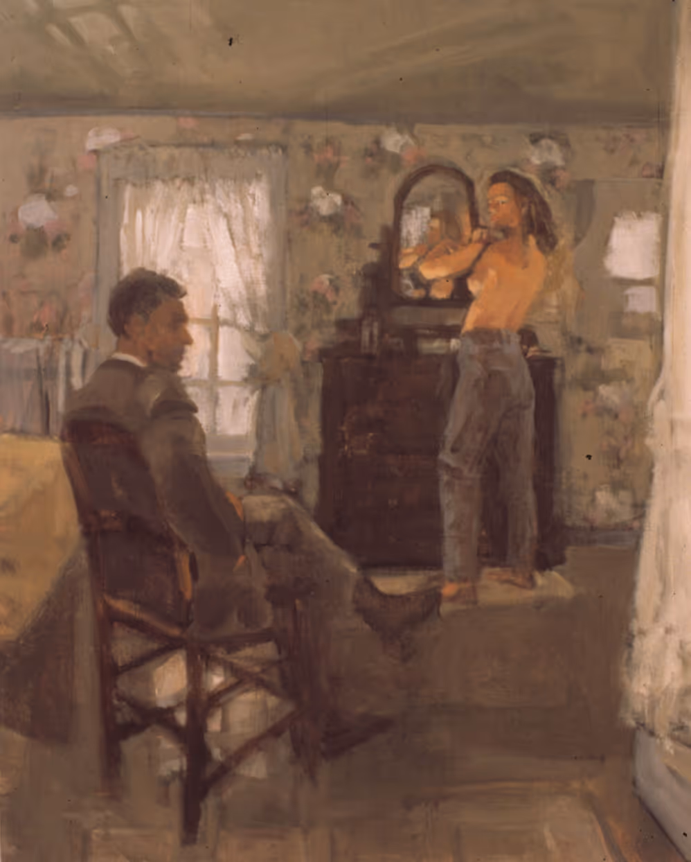 A man in a suit sits on a chair while a topless woman adjusts her hair near a dresser with a mirror.