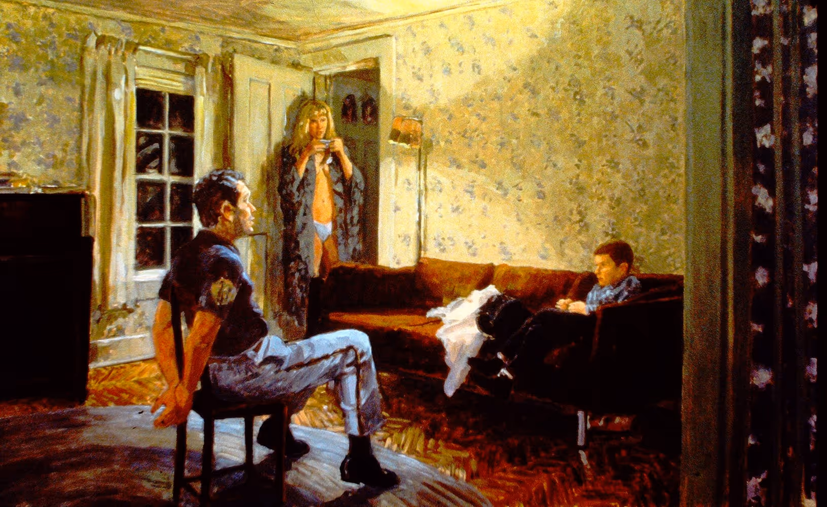 A boy sits on a sofa speaking to a man while his mother drinks from a cup in the doorway.