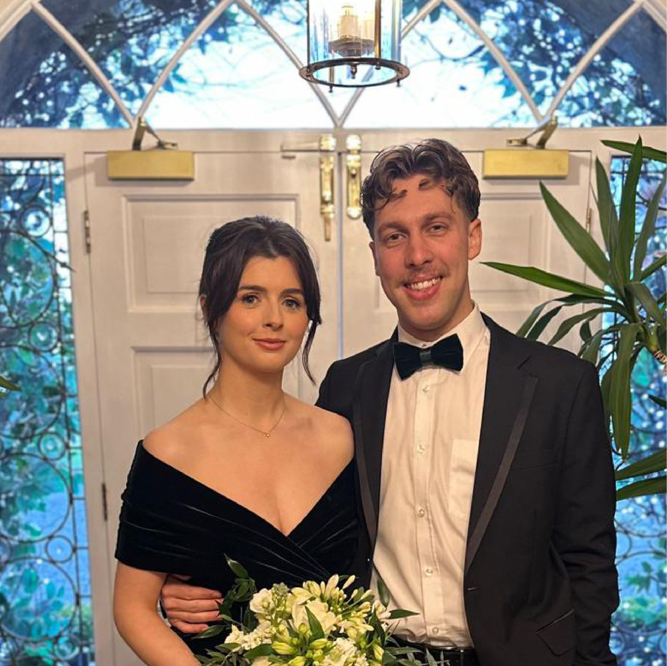 Man in tuxedo and woman in black dress holding bouquet in front of a white door with glass windows.