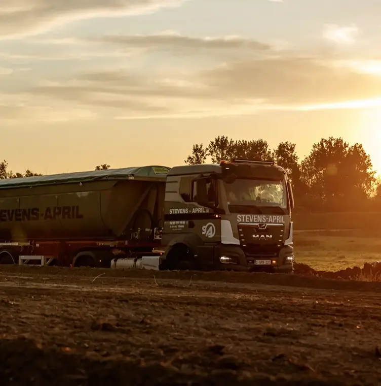 Vrachtwagen Stevens-April rijdt over modderig terrein bij zonsondergang met bomen op de achtergrond.