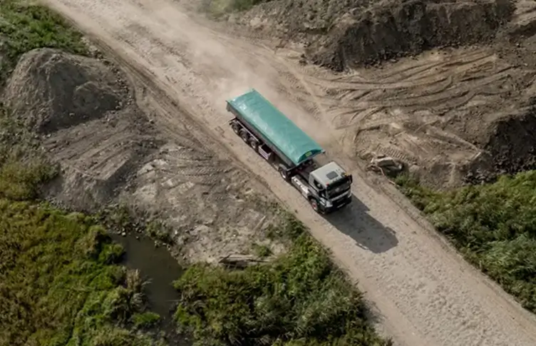 Vrachtwagen met groene afdekking rijdt over stoffige onverharde weg in een modderige werf.