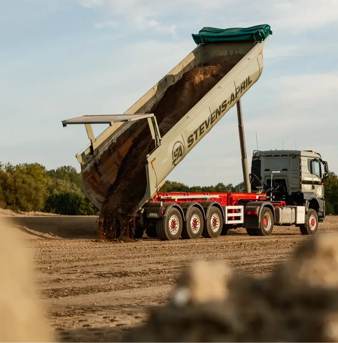 Kiepwagen aan het lossen van aarde op een bouwterrein met bomen op de achtergrond.