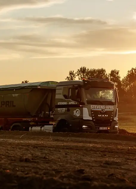Graafmachine rijdt op een bouwterrein bij zonsondergang met bomen op de achtergrond.