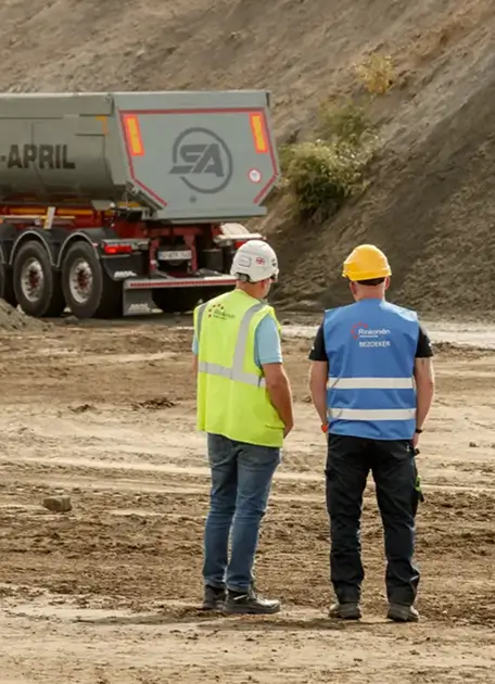 Twee bouwvakkers met helmen en vesten kijken naar een vrachtwagen op een bouwterrein.