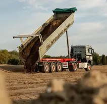 Kiepwagen kiept zand op een bouwterrein met bomen en lichte bewolking op de achtergrond.