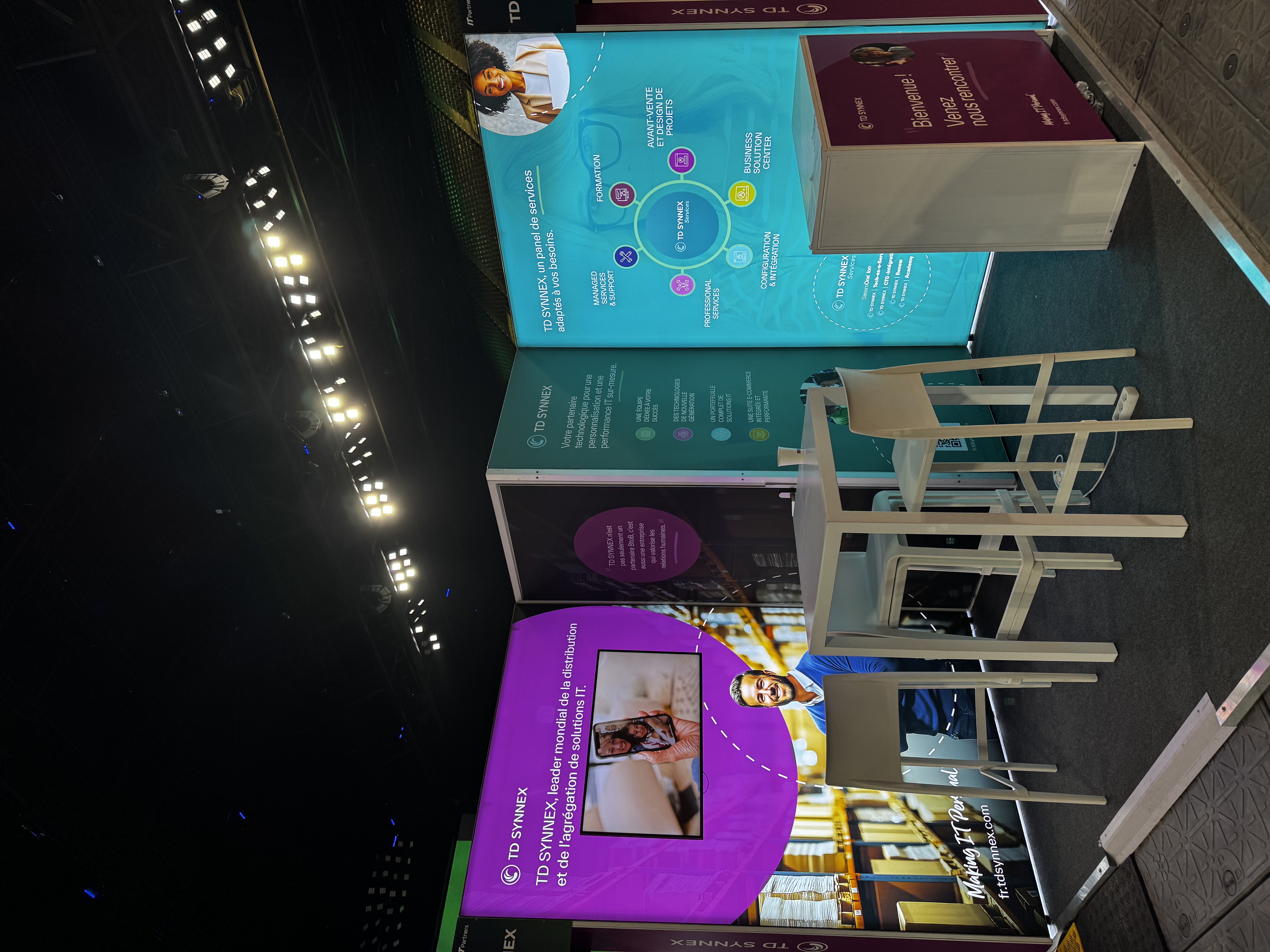 Trade show booth with bright purple and teal panels showcasing TD SYNNEX IT distribution and services, with a white table and chairs in front.