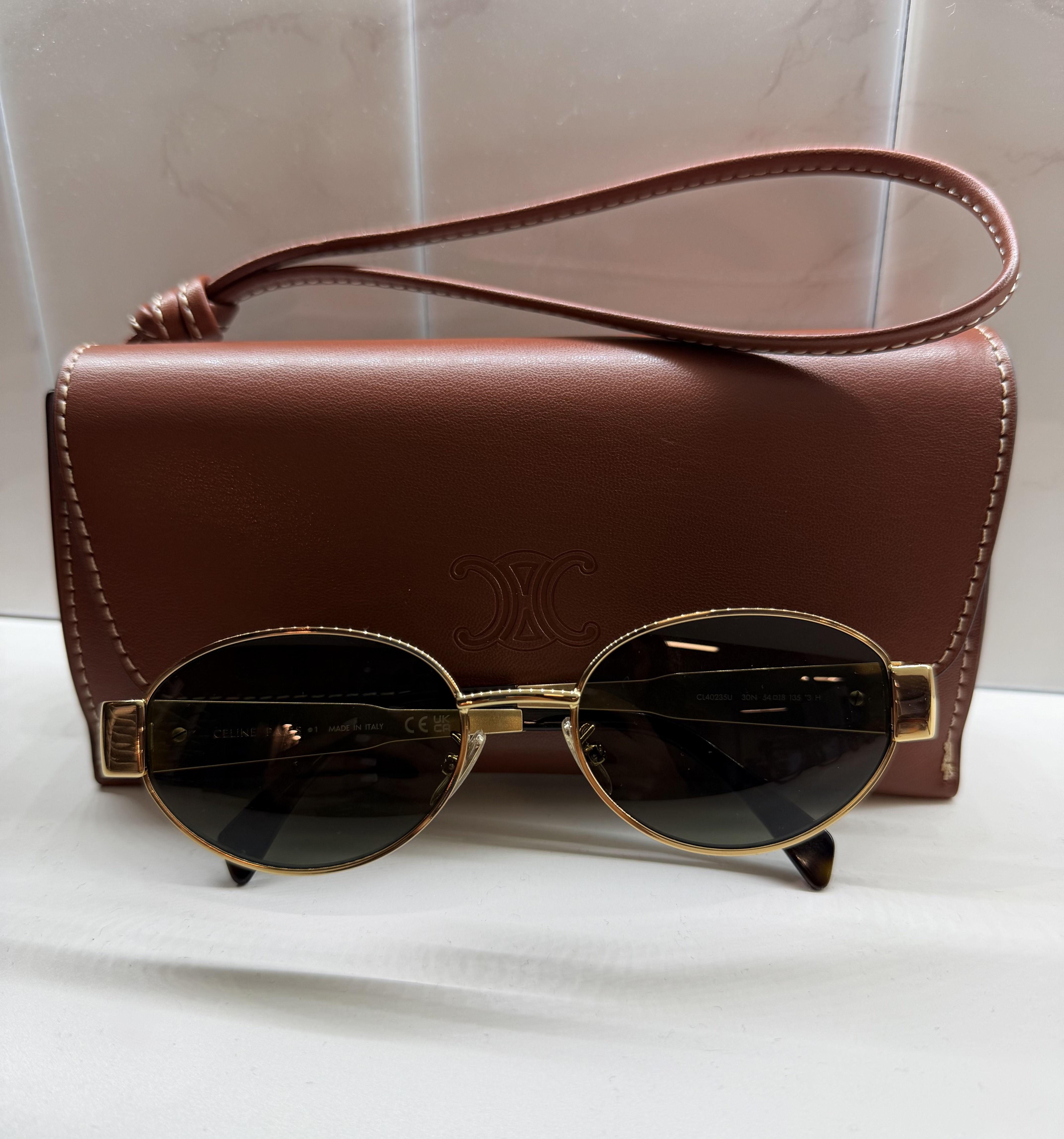 Brown leather shoulder bag with gold-framed oval sunglasses displayed in front