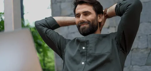 Bearded man in a green shirt leaning back with hands behind his head, smiling contentedly.