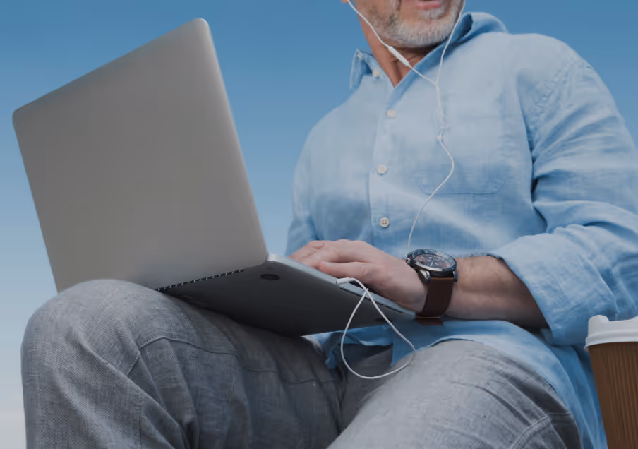 Man wearing a light blue shirt and gray pants using a laptop while seated, with earphones in and a coffee cup beside him.
