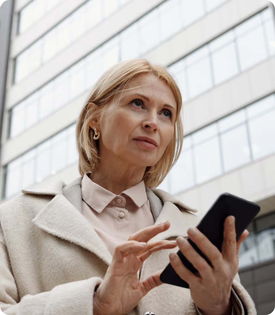 Middle-aged woman in a beige coat using a smartphone outdoors in front of a modern building.