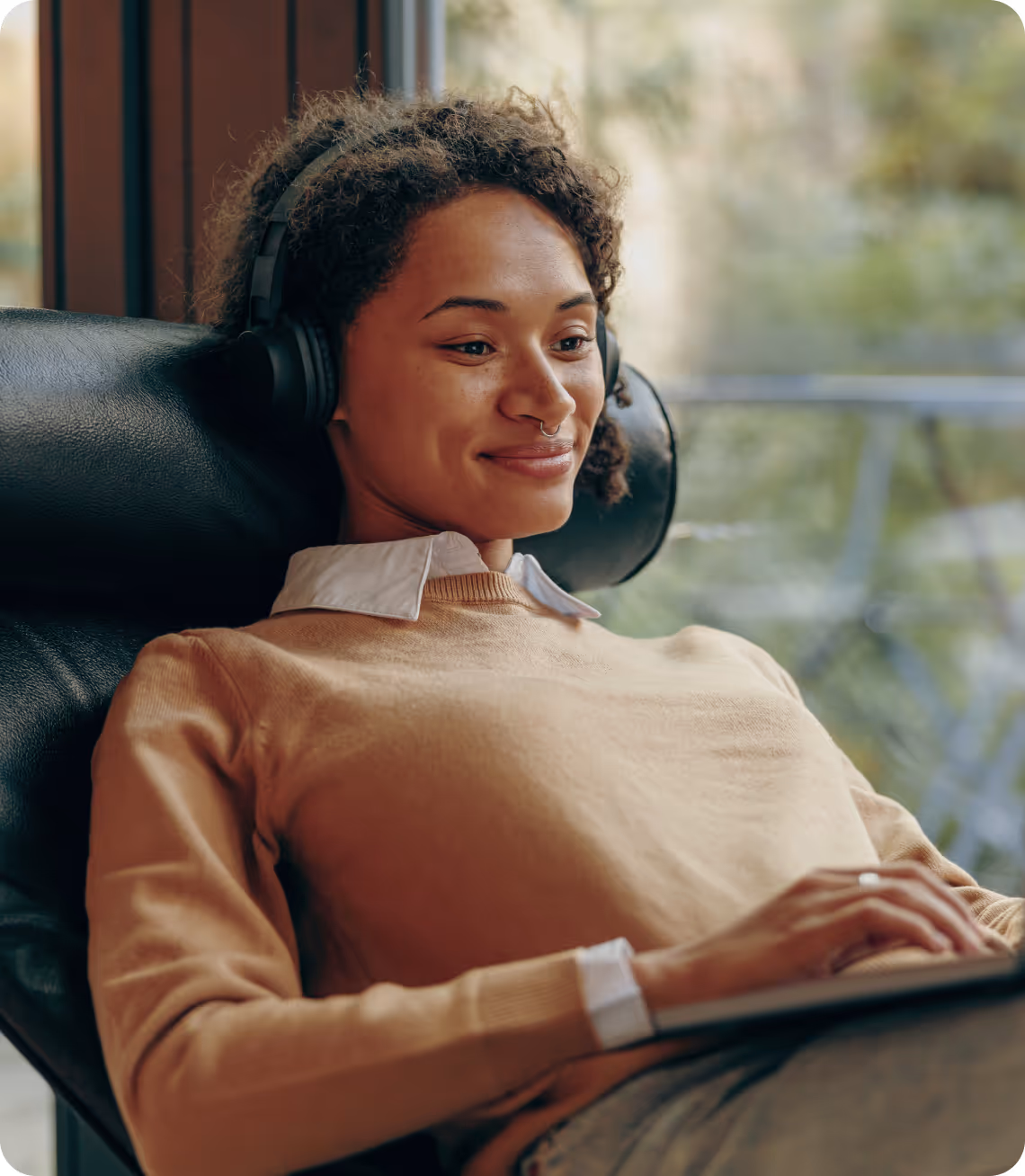 Young woman with curly hair wearing headphones and a beige sweater, smiling while using a laptop indoors.