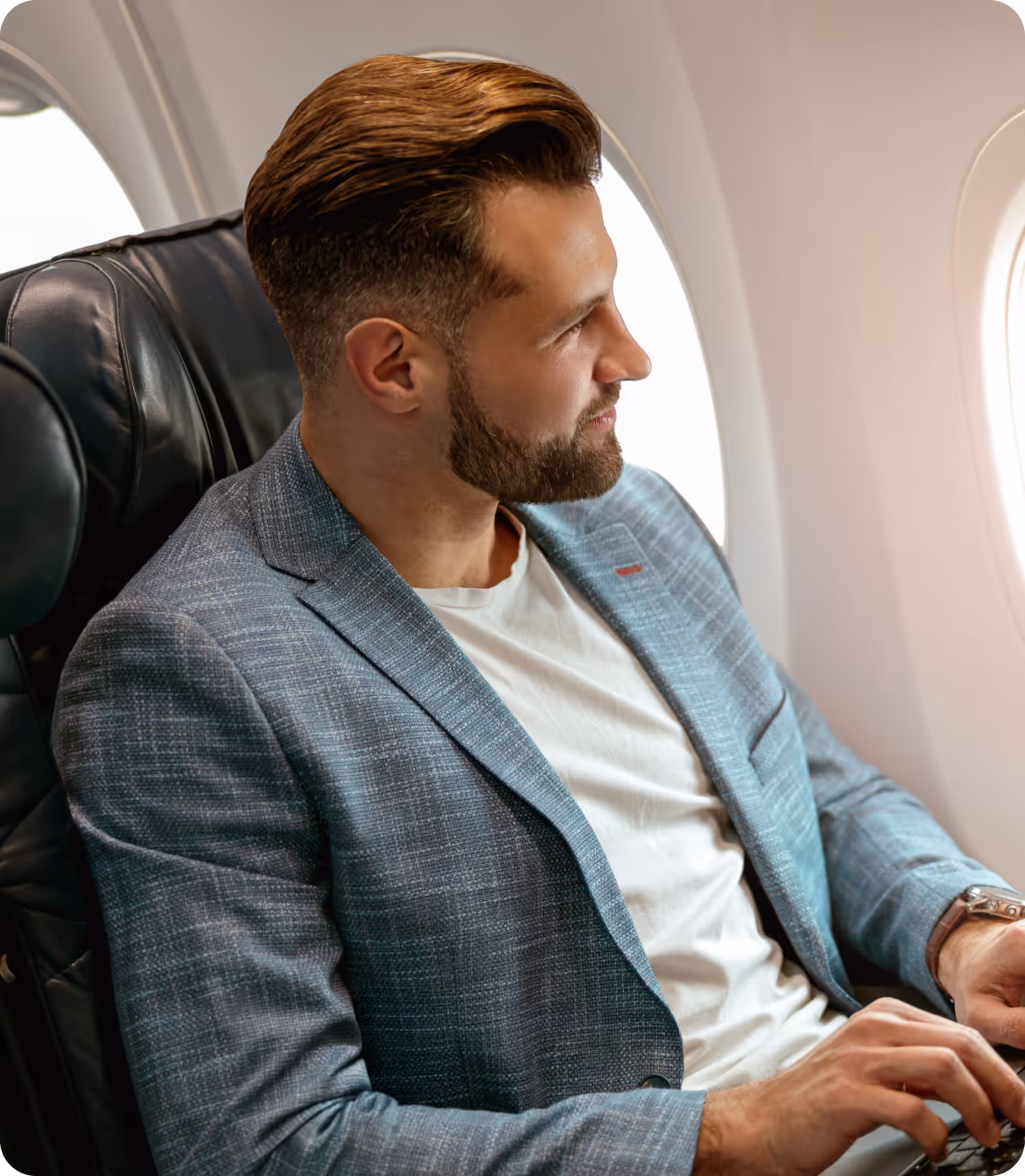 Man with neatly styled hair and beard sitting in an airplane seat working on a laptop.