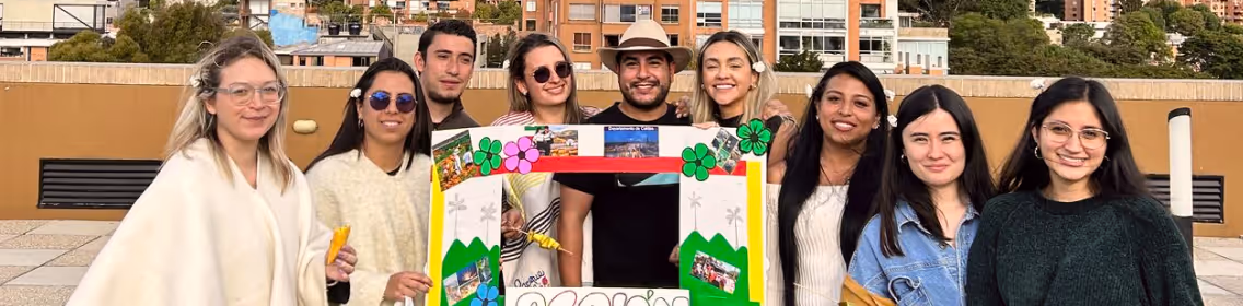 Group of nine people smiling behind a colorful decorated photo frame outdoors in an urban setting.