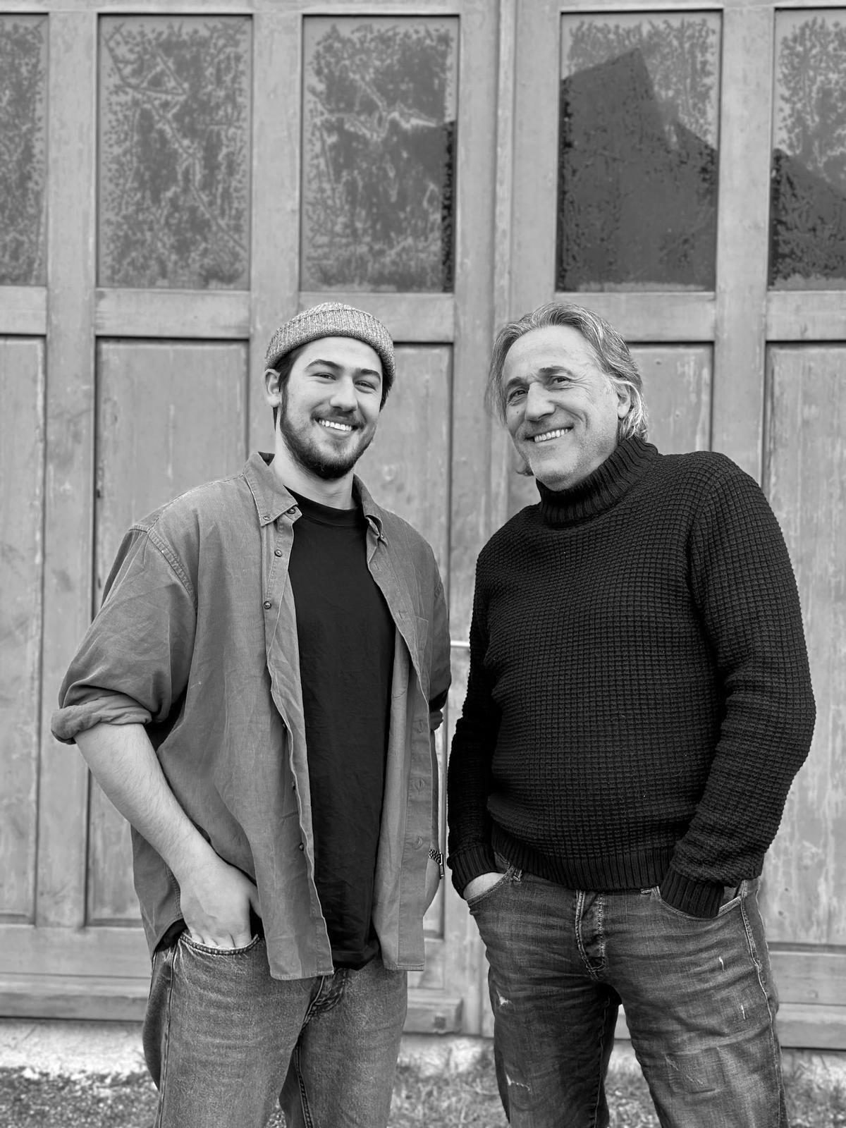 Zwei lächelnde Männer stehen vor einer Holzfassade, beide mit den Händen in den Taschen.