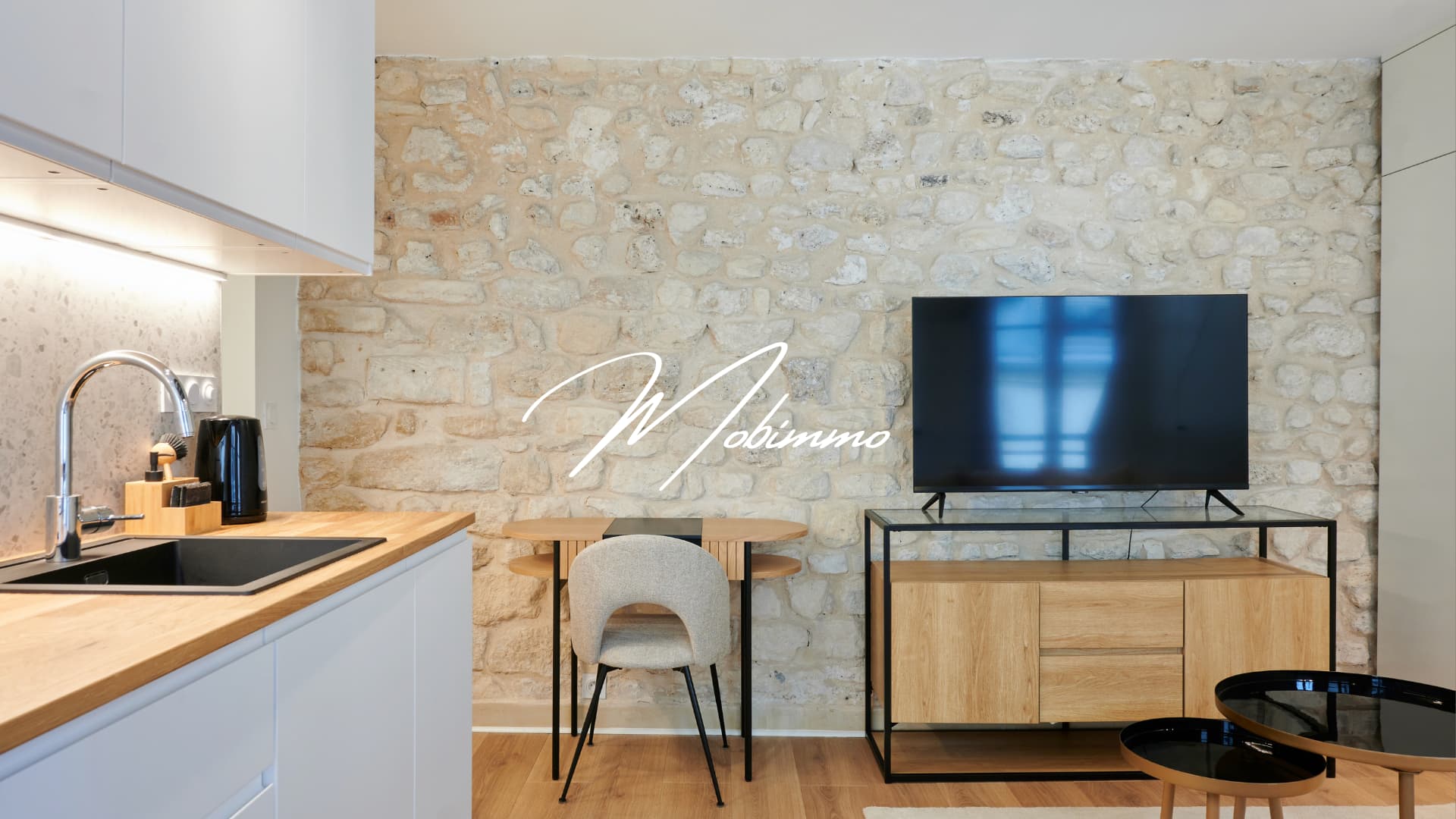 Salon T2 Amari Paris 6ème — ameublement LMNP, mur en pierre apparente et coin cuisine, Mobimmo