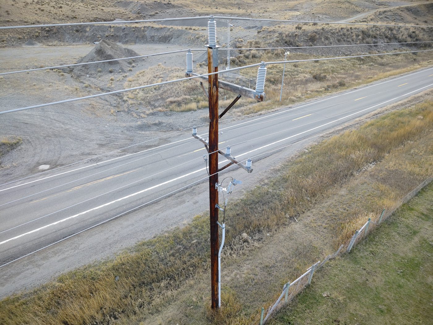 Aerial drone photograph of a wooden distribution pole with crossarms, insulators, and surge arresters beside a rural highway - the kind of asset a Part 2 shot sheet covers