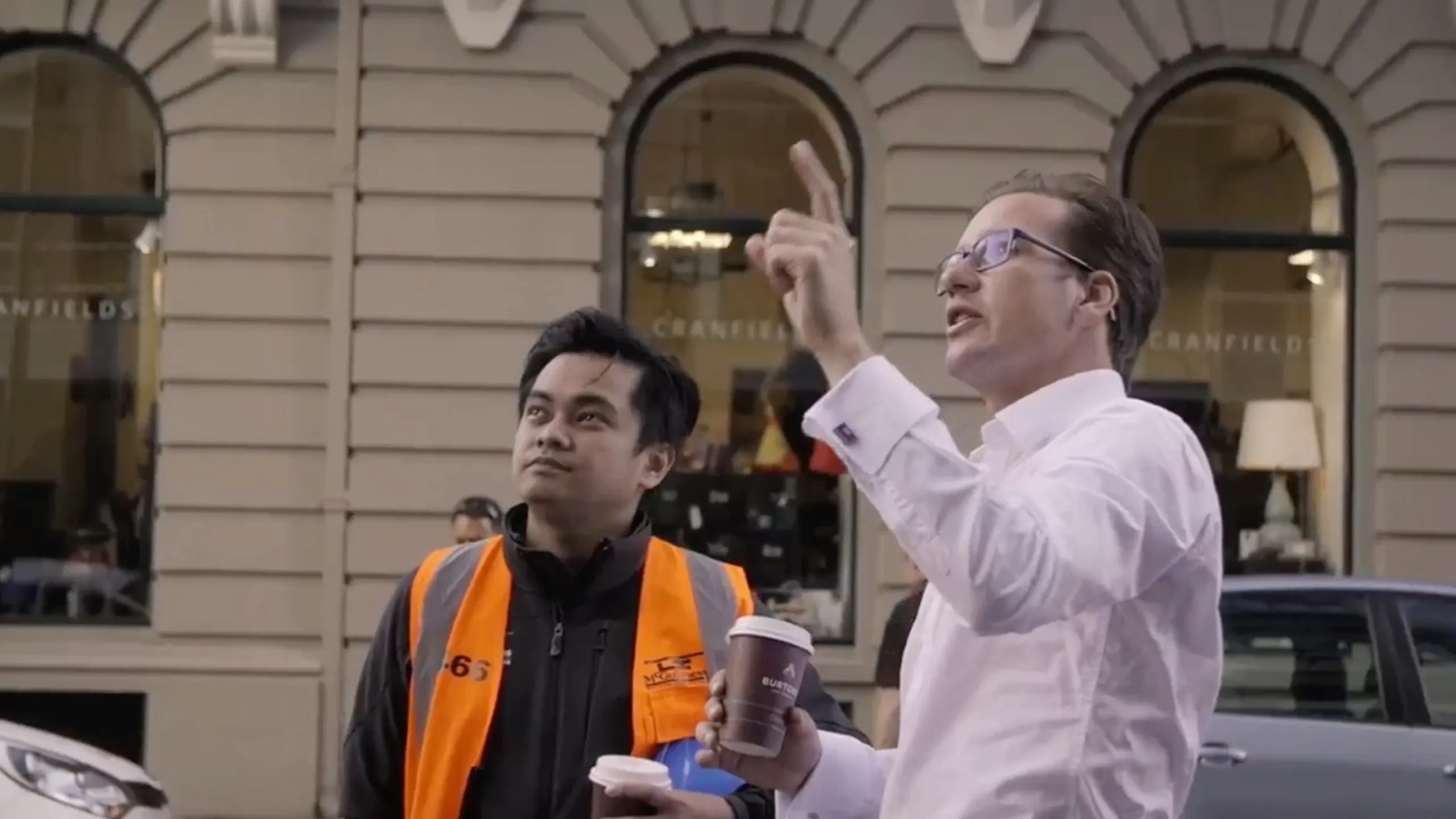 Two men holding coffee cups outside a building, one wearing an orange safety vest and the other in a white shirt pointing upwards.