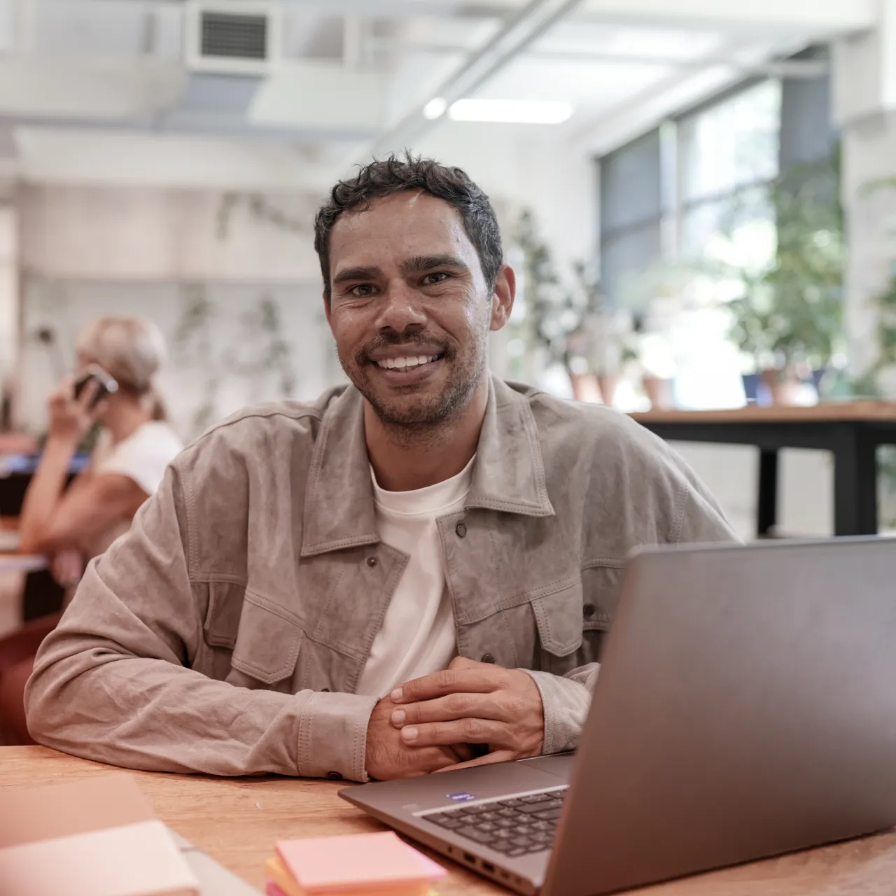 Smiling man with curly hair sitting at a desk with a laptop, in a bright office with plants and windows.